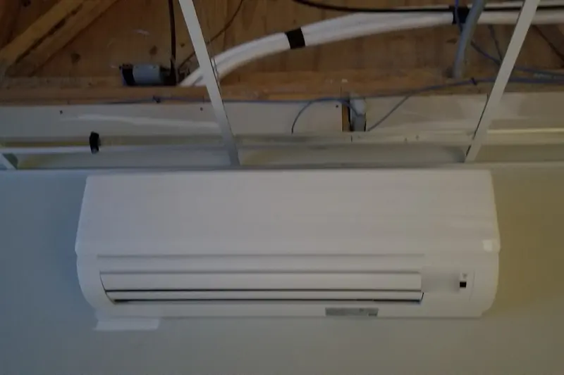 Photo of a white wall-mounted ductless head fixed high under exposed ceiling framing, with covered line sets nearby